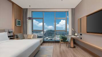 A modern hotel room with a large bed, a desk, a chair, a TV, and a balcony with a city view.