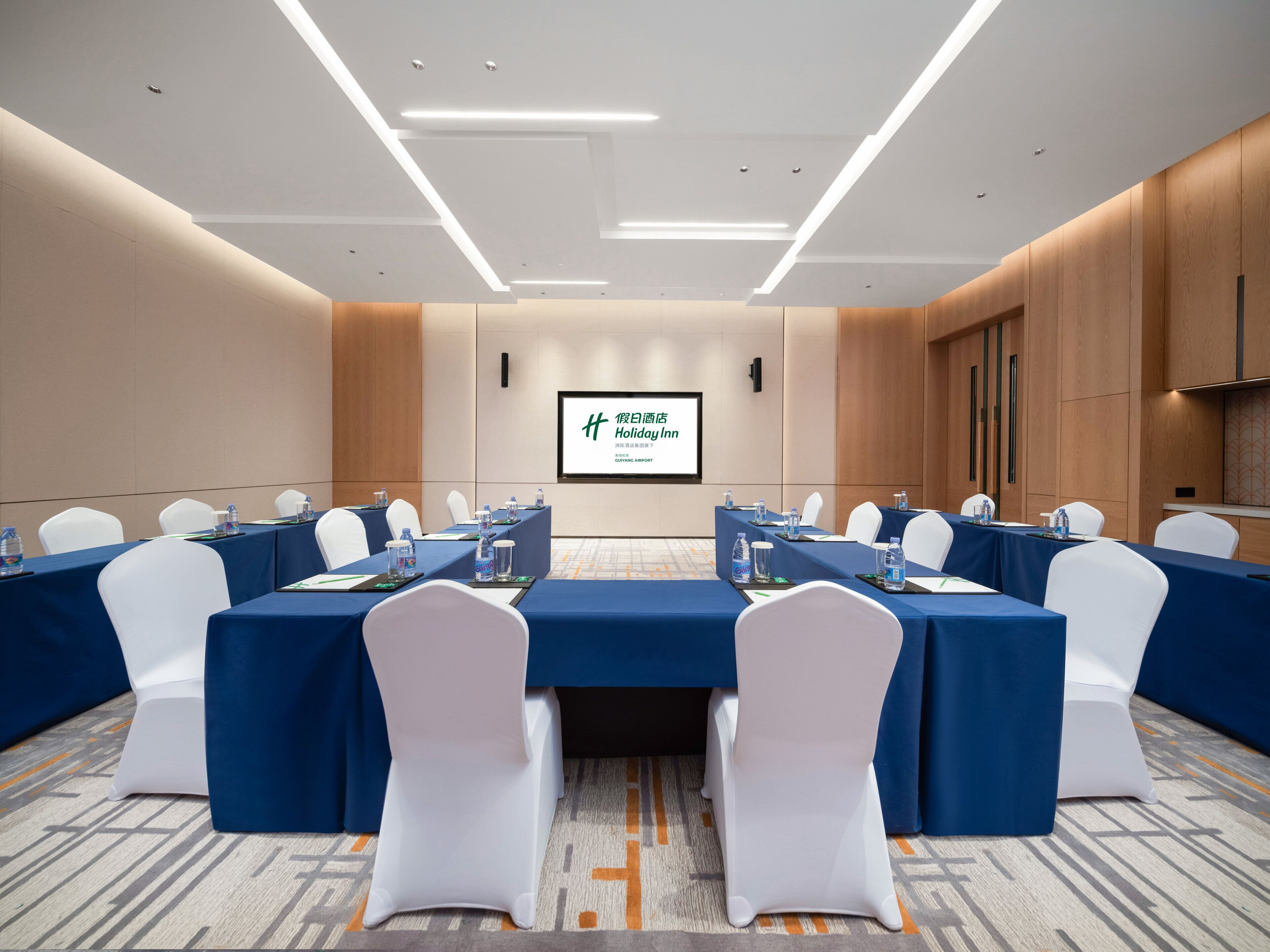 Un salón de conferencias con una larga mesa, sillas blancas y una pantalla que muestra información del hotel.