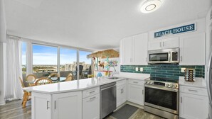 Una cocina moderna con armarios blancos, un horno de acero inoxidable, un microondas y una isla grande con un lavamanos. Un letrero sobre el microondas dice 'BEACH HOUSE'.