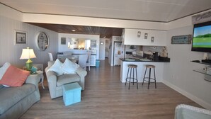 Ein modernes Wohnzimmer mit einer Couch, einem Couchtisch und einem Fernseher. Es gibt eine Küchenzeile mit einer Bar und Hockern.