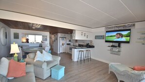 Ein modernes Wohnzimmer mit Küche im Hintergrund, einem an der Wand befestigten Fernseher und einem kleinen Essbereich.