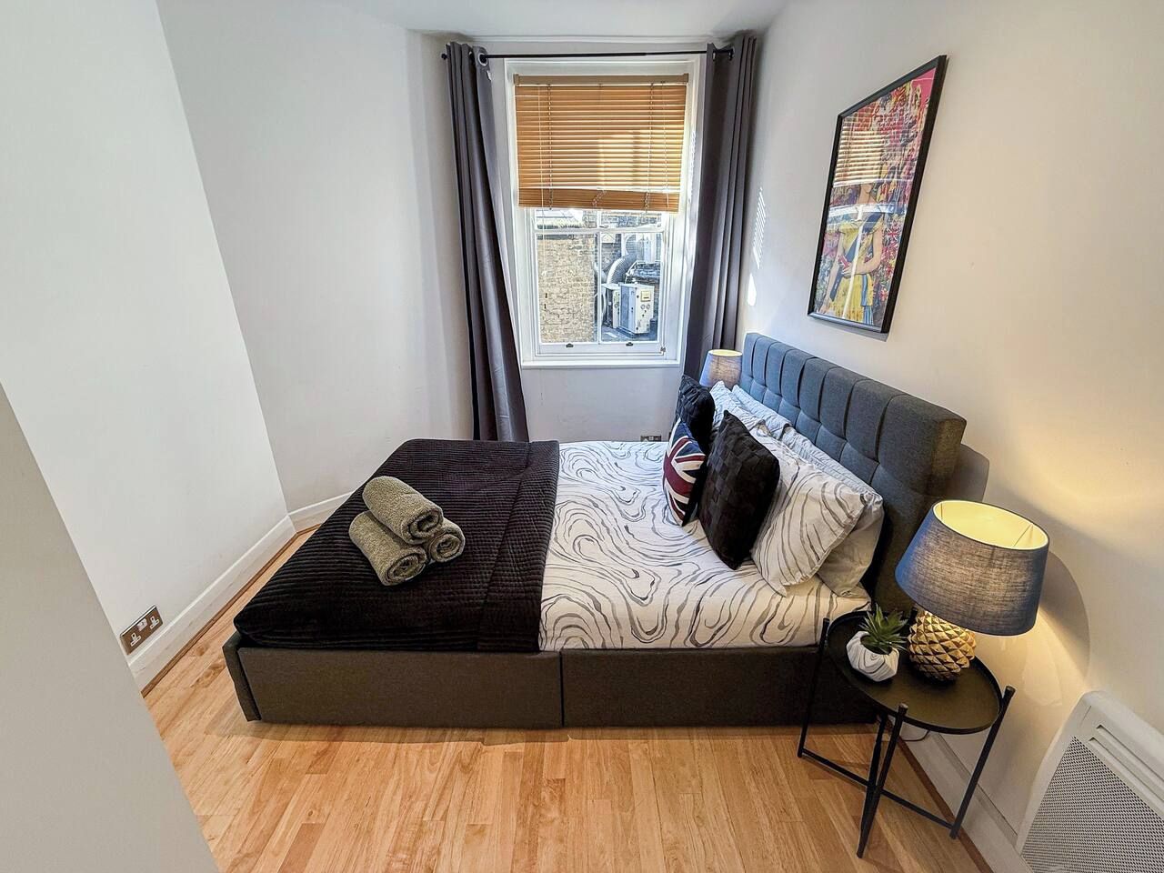 Une chambre à coucher avec un lit, une fenêtre avec des rideaux, une œuvre d’art accrochée au mur, une lampe de chevet et une petite table sur laquelle est posée une plante.