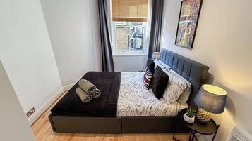 Une chambre à coucher avec un lit, une fenêtre avec des rideaux, une œuvre d’art accrochée au mur, une lampe de chevet et une petite table sur laquelle est posée une plante.