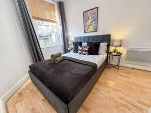 A bedroom with a bed, a window, a bedside table with a lamp, and a wall art piece.