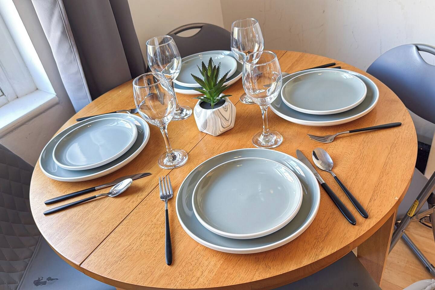 A round wooden dining table set with plates, cutlery, and wine glasses, with a small potted plant in the center.