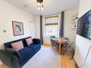 A living room with a blue sofa, a dining table with chairs, a TV, and a window with curtains.