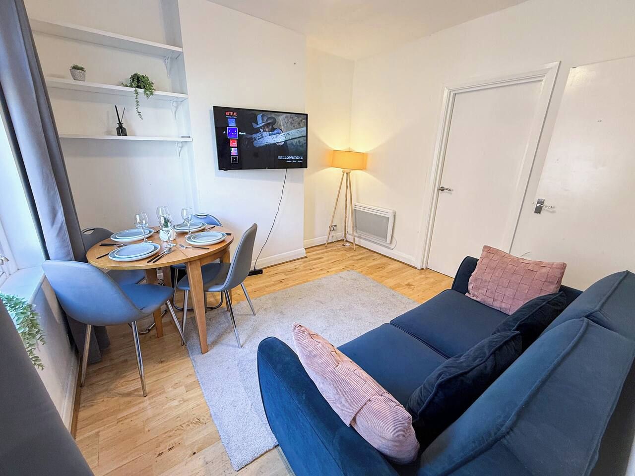 A living room with a blue sofa, a dining table with chairs, a TV mounted on the wall, and a lamp.