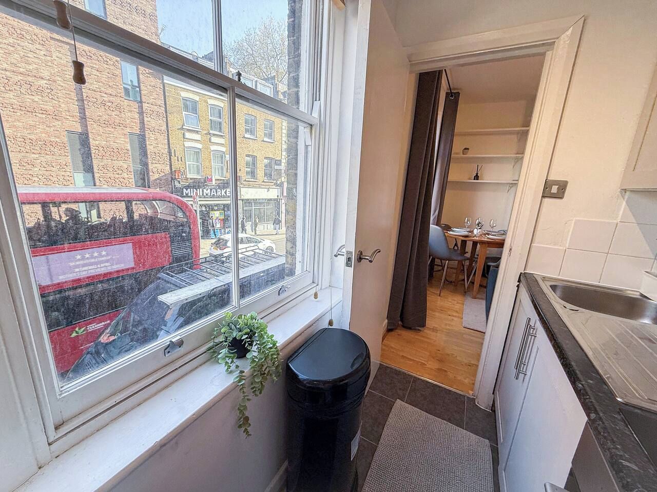 A kitchen with a window, a trash bin, and a view of a street with a red bus.