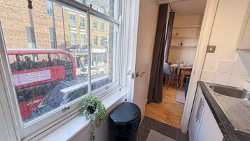 A kitchen with a window, a trash bin, and a view of a street with a red bus.