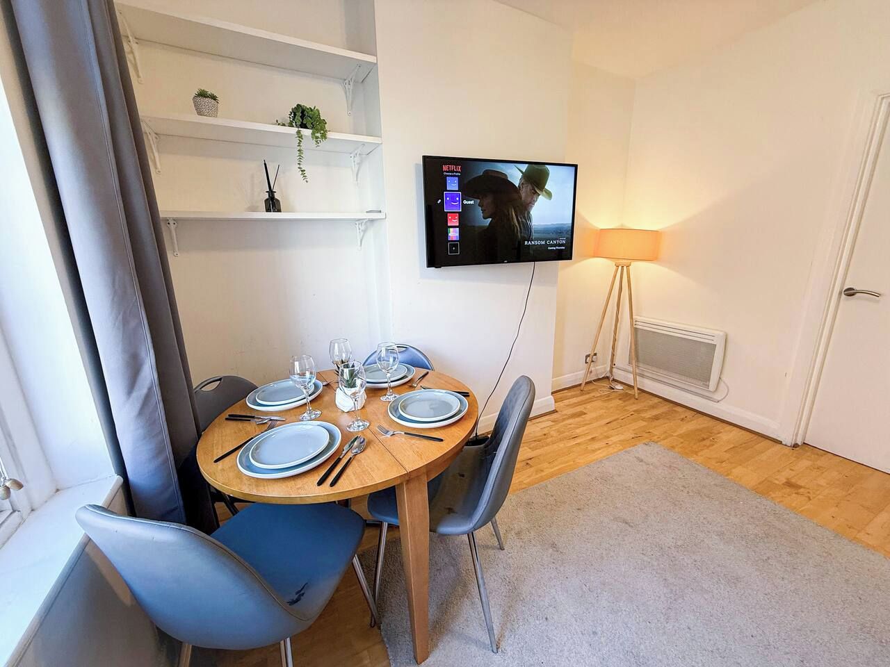 Ein Essbereich mit einem runden Tisch, gedeckt für vier Personen, einem an der Wand montierten Fernseher und einer Stehlampe.
