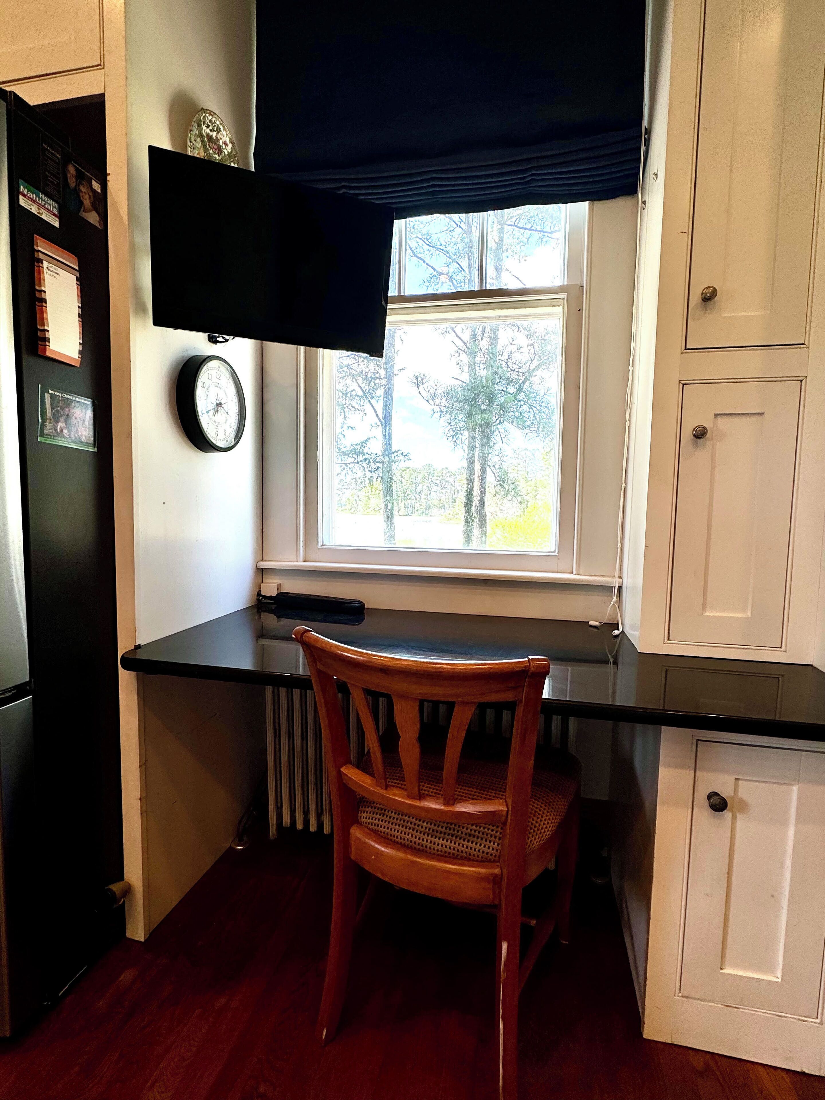 Una cocina con una silla de madera, una encimera negra, una ventana con vistas a árboles y un reloj en la pared.