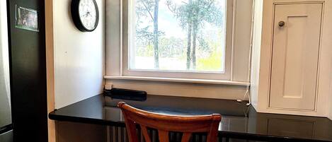 Una cocina con una silla de madera, una encimera negra, una ventana con vistas a árboles y un reloj en la pared.