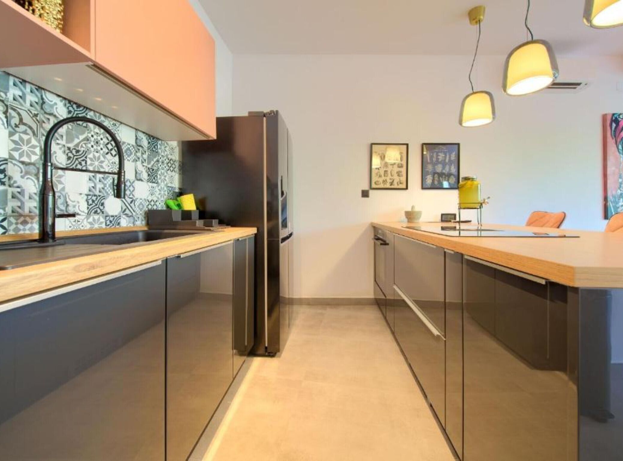 A modern kitchen with a stainless steel refrigerator, a wooden countertop, and a backsplash with a geometric pattern. There are two pendant lights above the counter and a small table with a yellow lamp.
