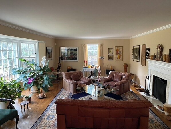 A living room with two leather sofas, a glass coffee table, and a fireplace. There are potted plants and artwork on the walls.
