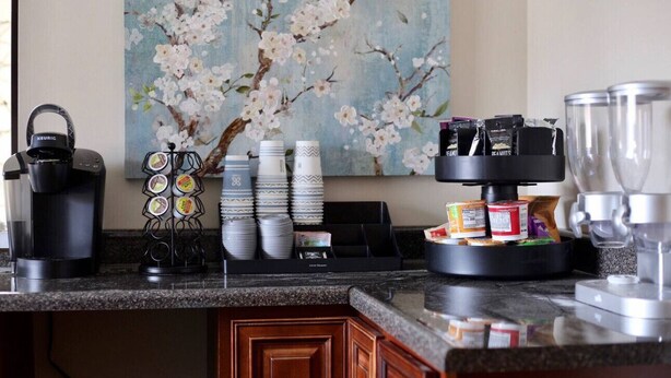 A kitchen counter with a coffee maker, a stack of plates, a coffee dispenser, and a food display.