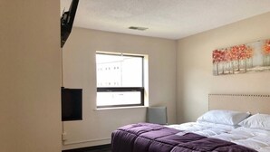 A bedroom with a bed, a window, a painting, and a wall-mounted TV.