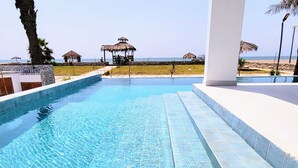 A swimming pool with clear blue water, a white tiled deck, and a view of the beach with thatched-roof structures. - Hermosa Casa Frente AL MAR CON Piscina Privada (low chincha)