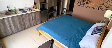 A compact hotel room with a bed, a small desk, a chair, a kitchenette, and a bathroom.