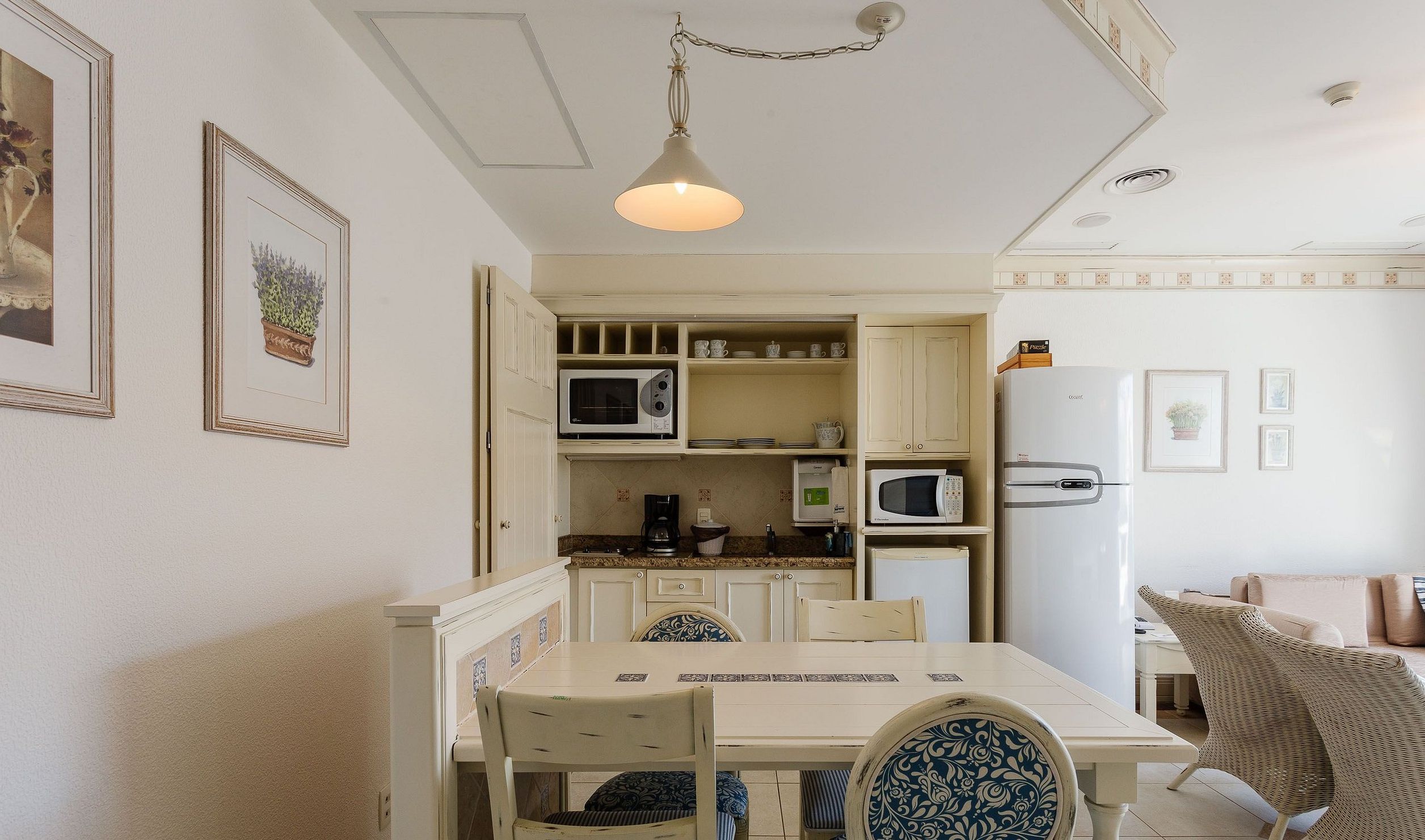 A compact kitchen with white cabinetry, a microwave, a coffee maker, and a refrigerator. There is a dining table with chairs, and the room has a ceiling light fixture.