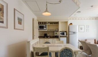 Cocina compacta con armarios blancos, microondas, cafetera y refrigerador. Hay una mesa de comedor con sillas, y la habitación tiene una lámpara en el techo.
