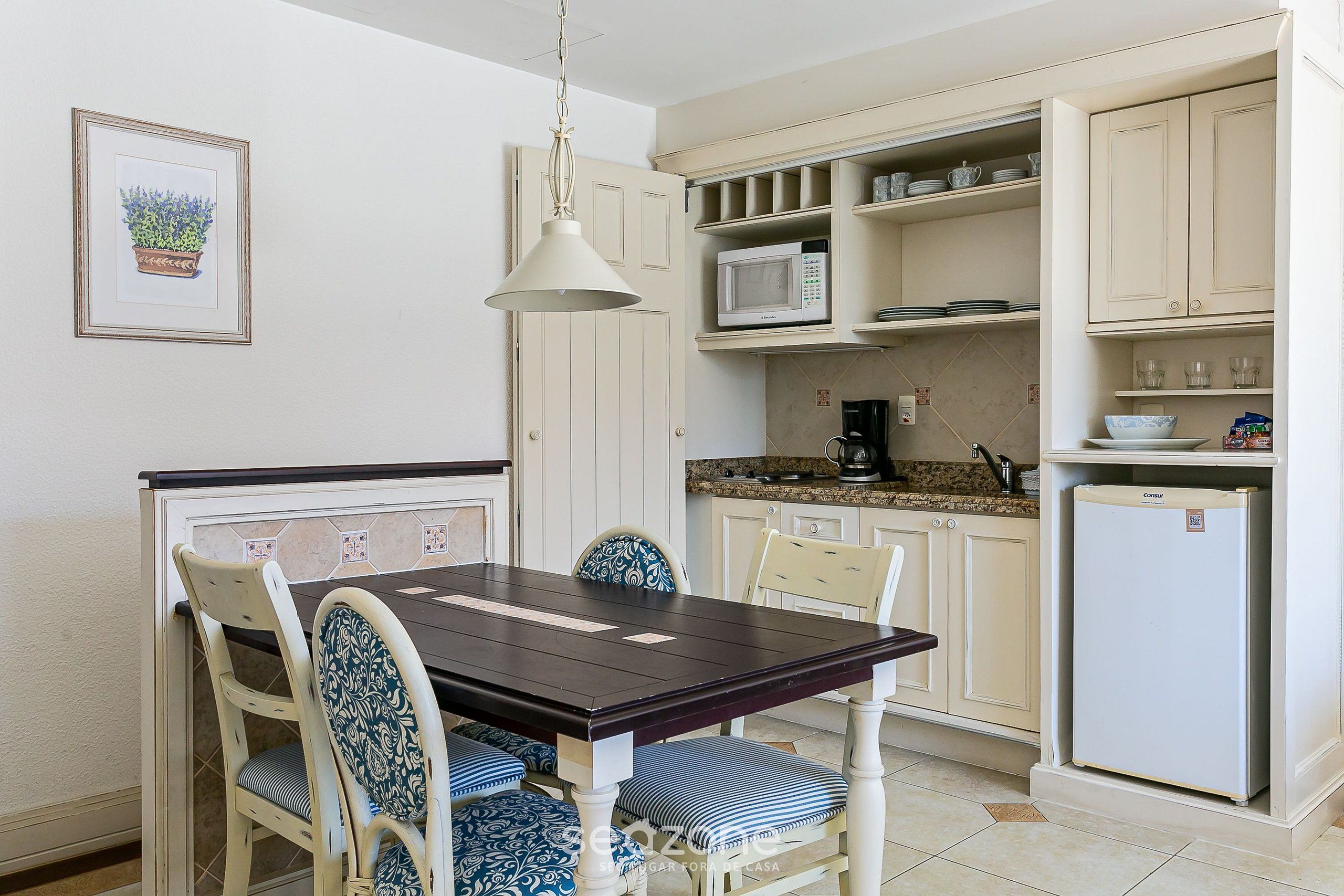 Un comedor con mesa y sillas de madera, una lámpara colgante y una cocina con muebles y electrodomésticos blancos.