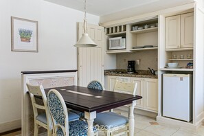 Un comedor con mesa y sillas de madera, una lámpara colgante y una cocina con muebles y electrodomésticos blancos.