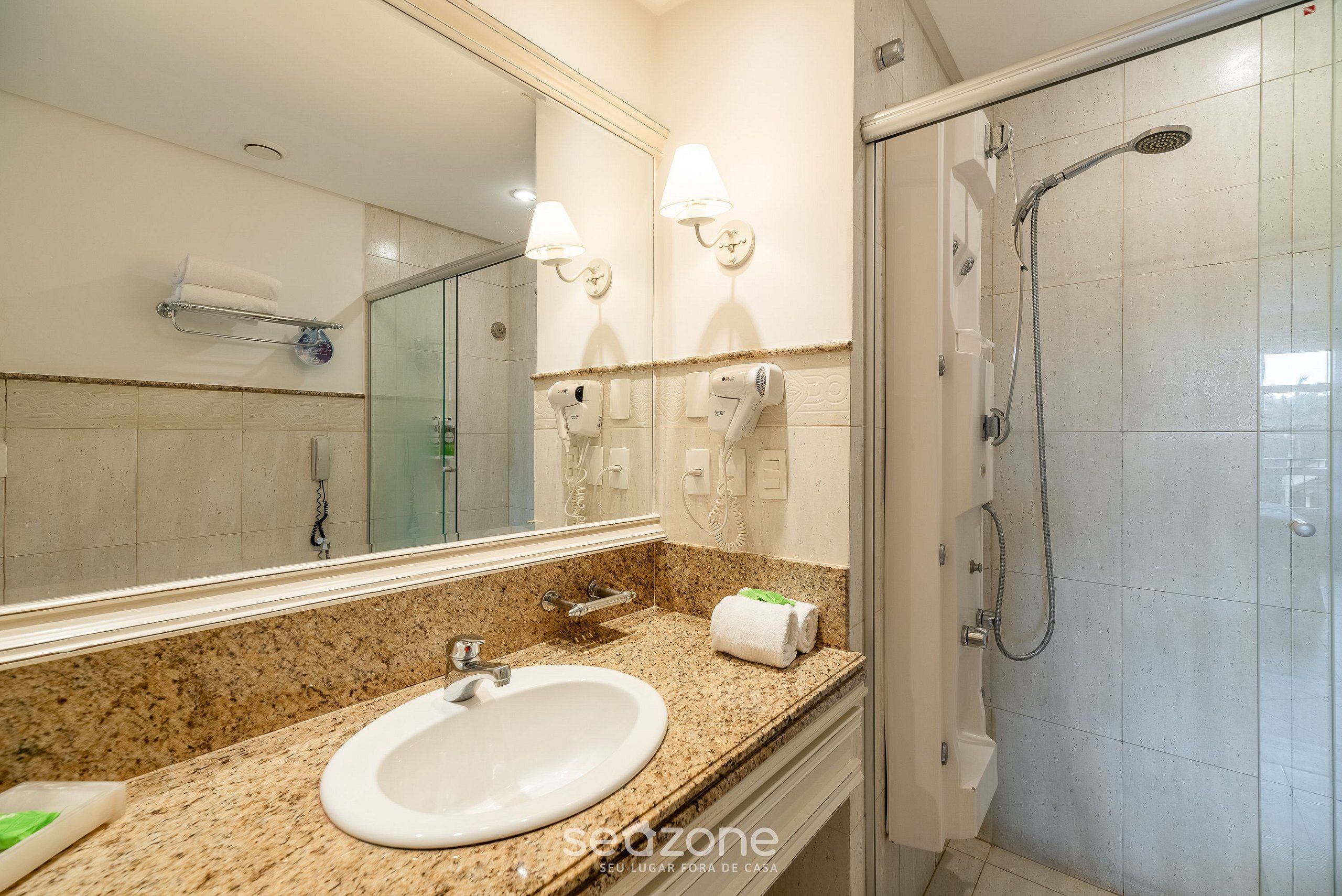 A bathroom with a granite countertop sink, a large mirror, a handheld showerhead, and a towel rack.