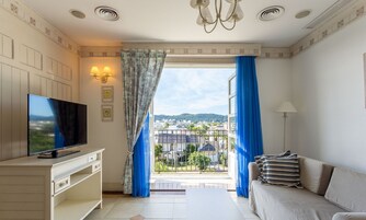 Una sala de estar con televisor, sofá y vista a una zona residencial a través de una ventana con cortinas azules.