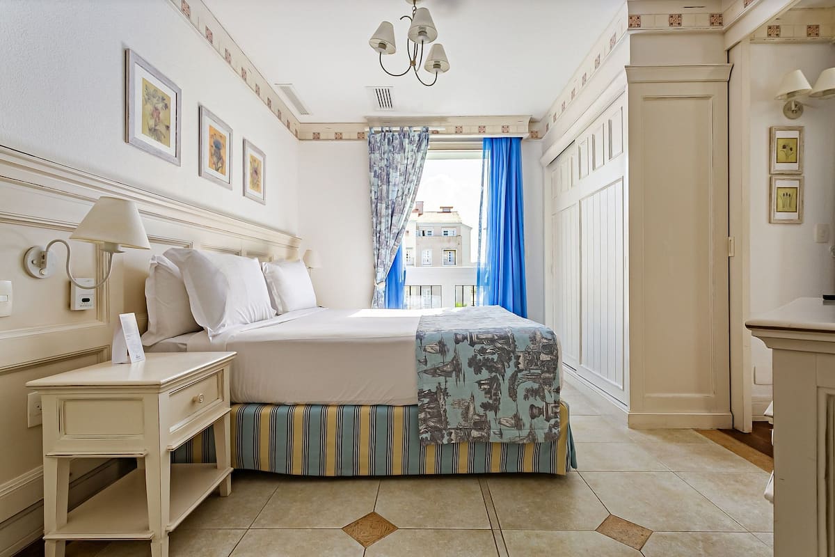 Habitación de hotel con cama, mesita de noche, lámpara, ventana con cortinas azules y vistas a edificios.
