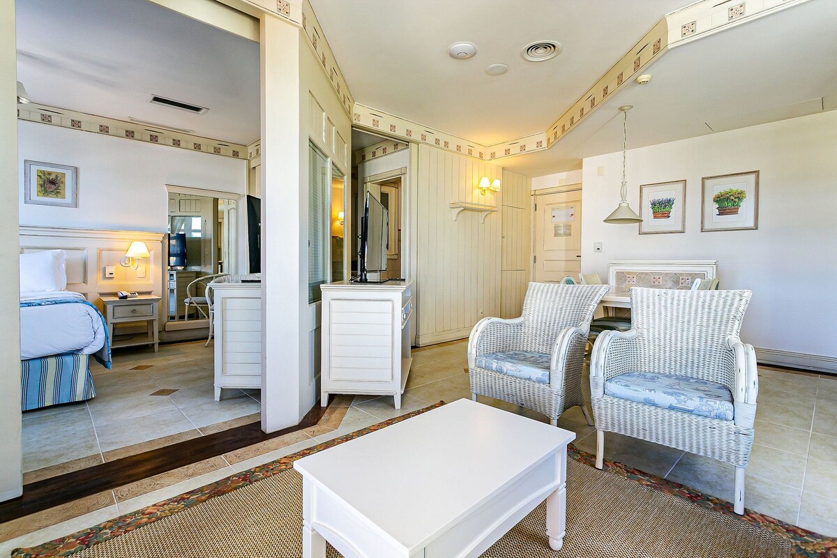 A hotel room with a bed, a white dresser, two wicker chairs, a white coffee table, a mirror, and a small dining area.