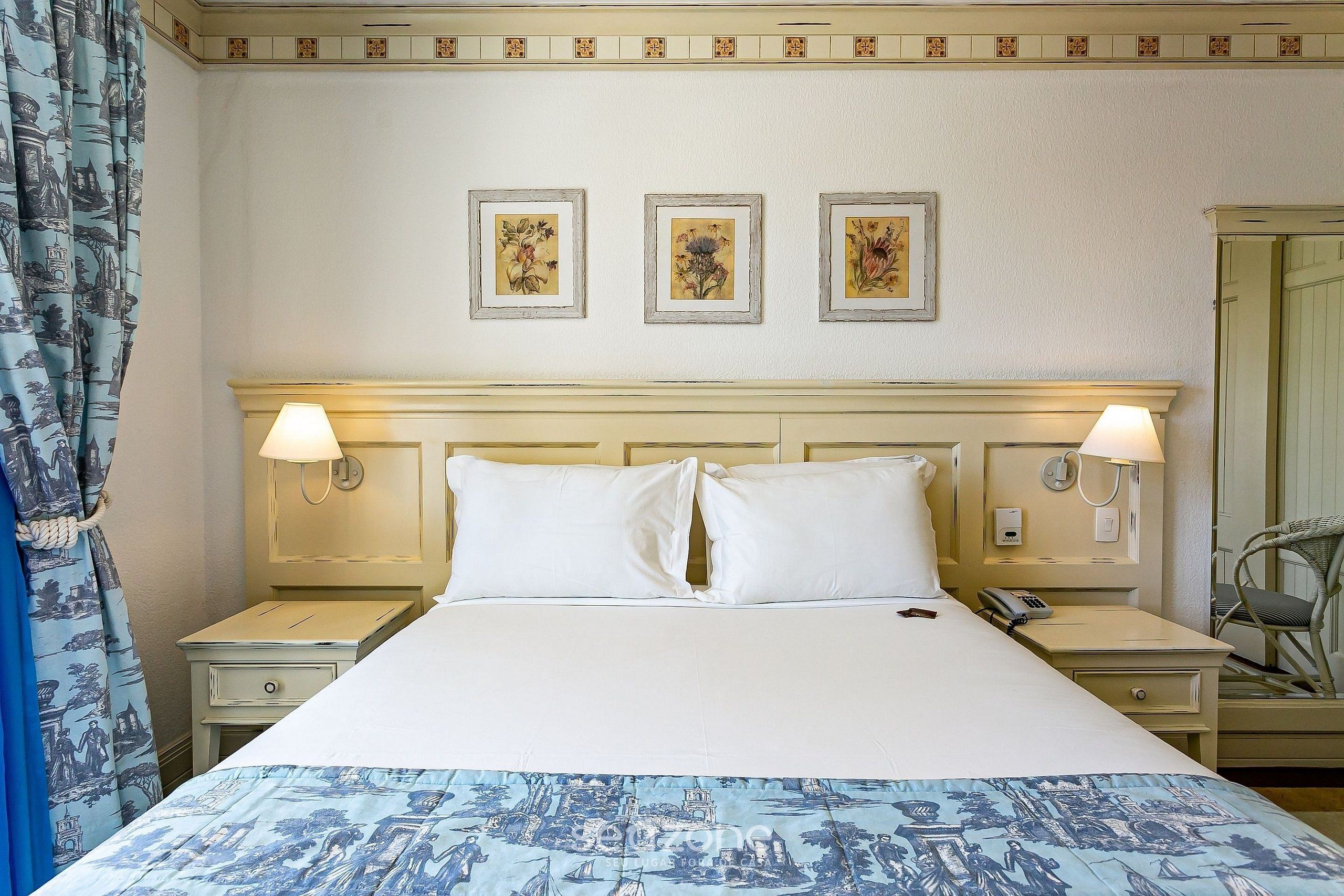 Une chambre d’hôtel comprenant un lit, deux tables de chevet, une petite table, une lampe, un téléphone et trois tableaux encadrés accrochés au mur.