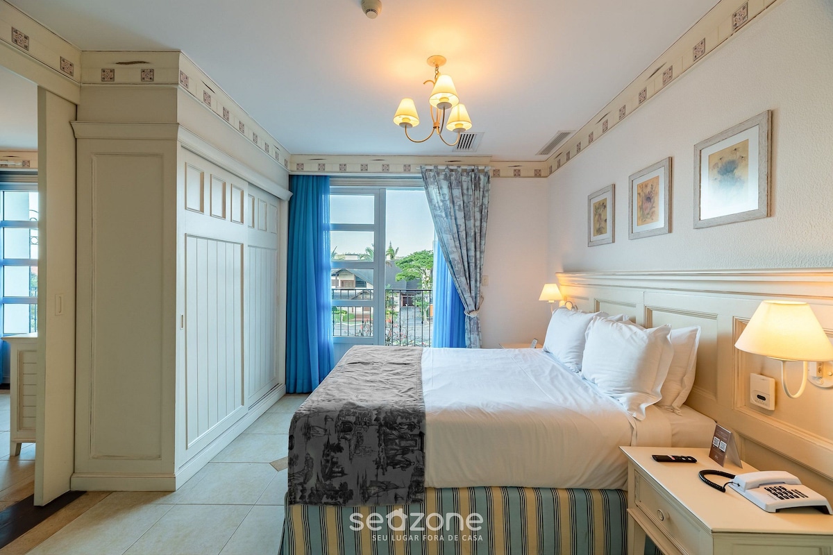 Habitación de hotel con una cama grande, una mesita de noche con lámpara y vistas al exterior a través de una ventana con cortinas azules.