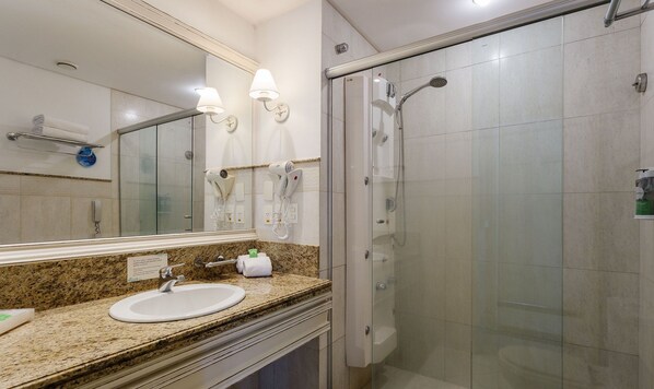 A bathroom with a glass-enclosed shower, a sink with a granite countertop, and a mirror with wall-mounted lights. - Studios frente ao mar no IL Campanario Resort ILCTOP (Florianópolis)