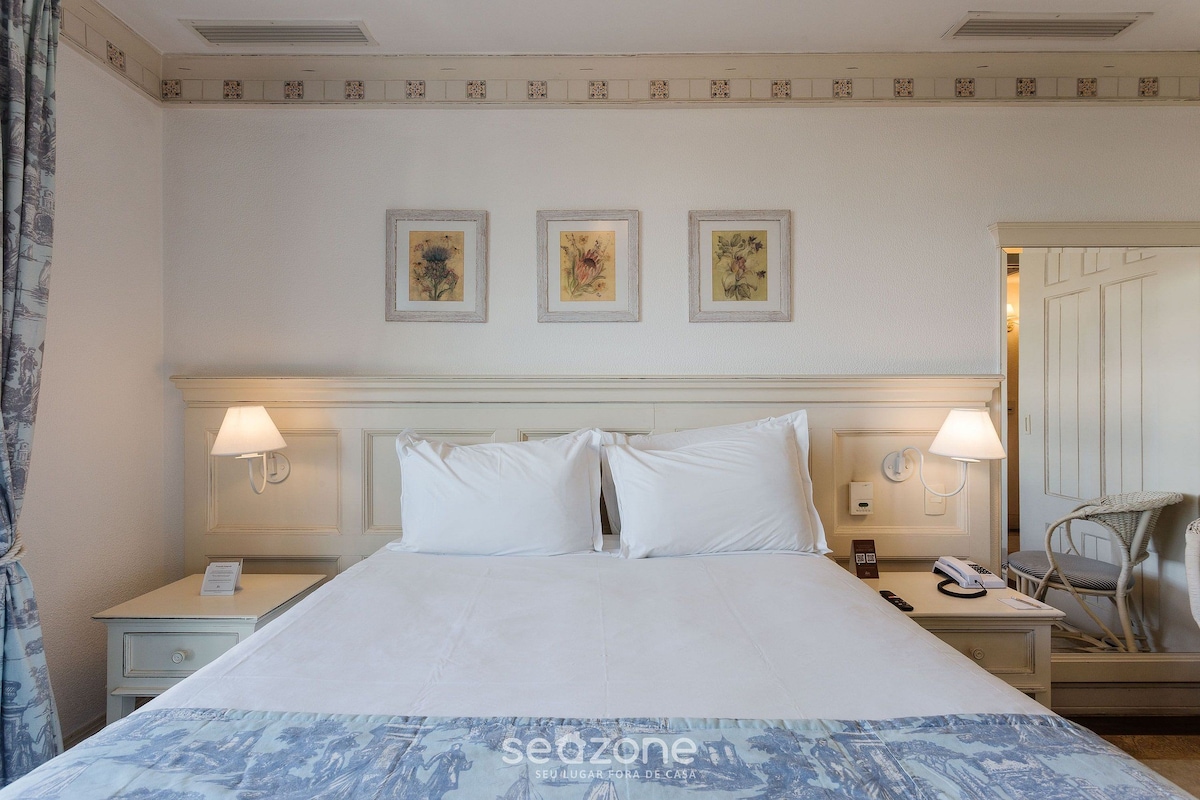 A hotel room with a large bed, two bedside tables, a chair, and three framed pictures on the wall.