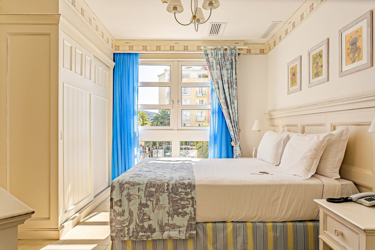 A hotel room with a bed, a bedside table, a lamp, and a window with blue curtains.