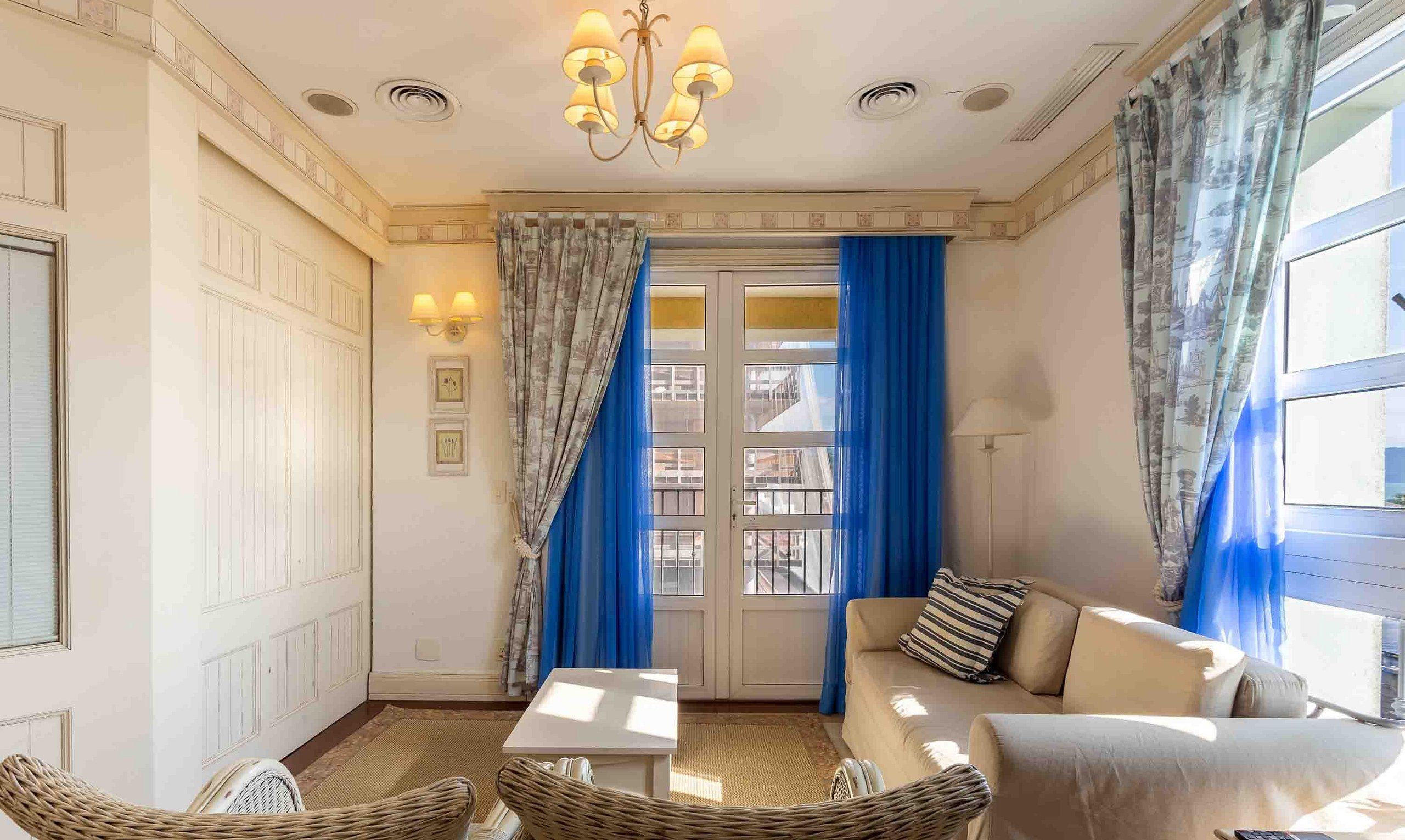 A room with a sofa, two wicker chairs, a small table, a chandelier, and a view through a window with blue curtains.