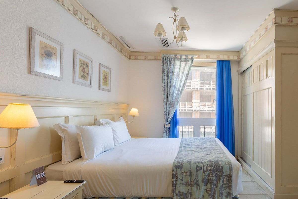 Habitación de hotel con cama, dos lámparas de noche, ventana con cortinas azules y vista a edificios.