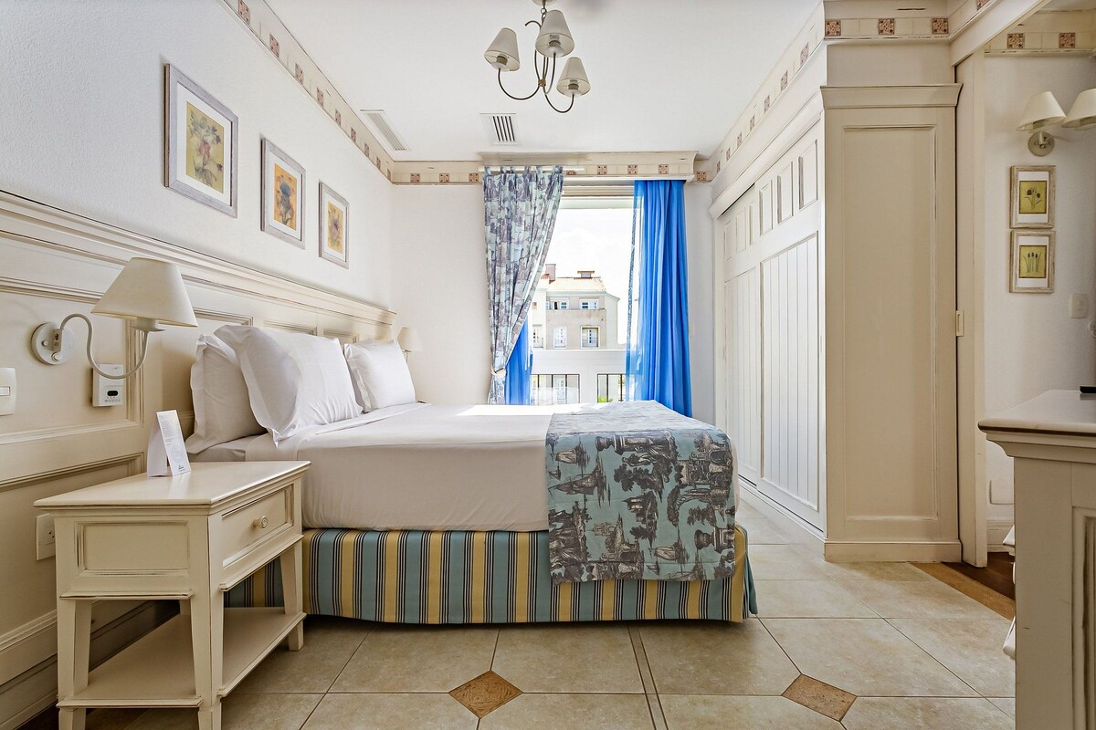 A hotel room with a bed, a nightstand, a lamp, a window with blue curtains, and a view of buildings outside.