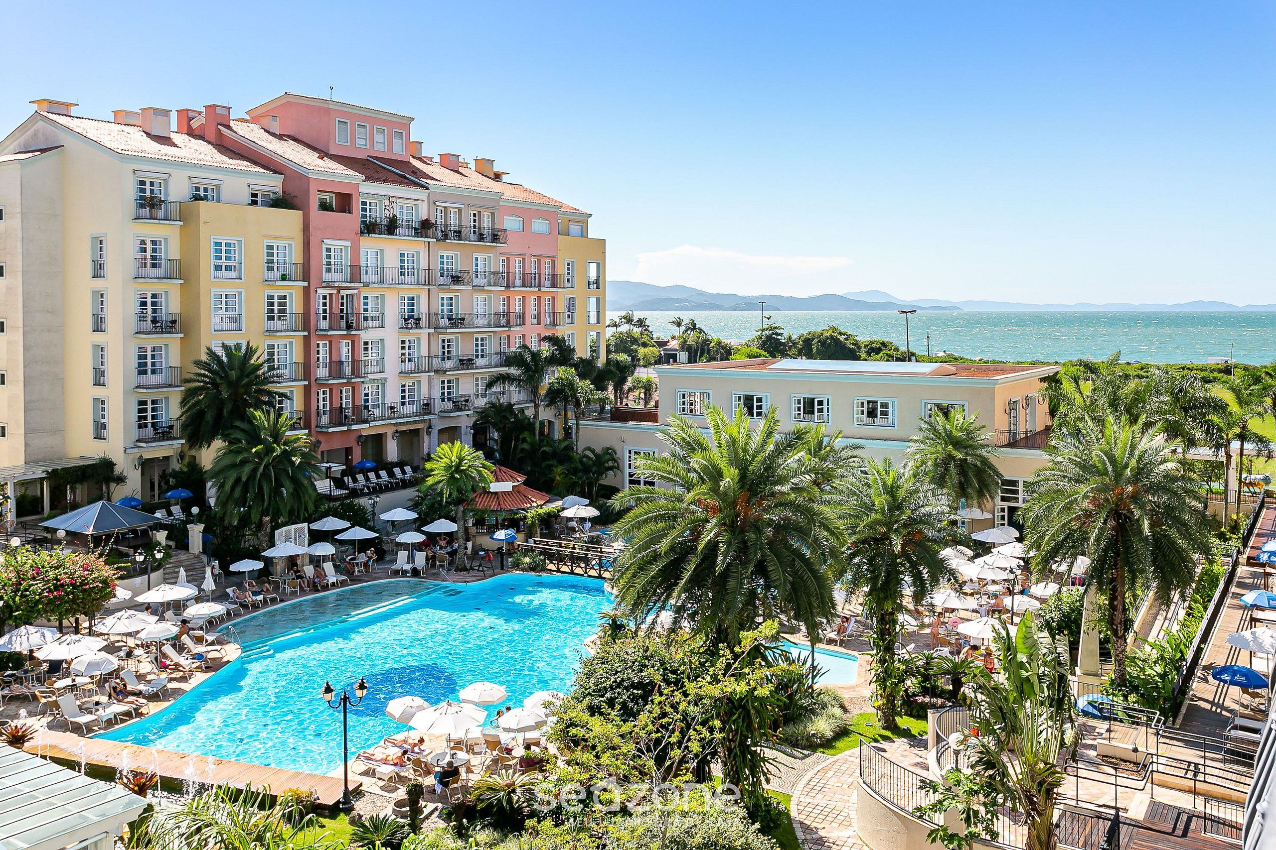 Ein Hotel mit Pool, umgeben von Palmen und Sonnenschirmen, mit Blick auf ein Gewässer.