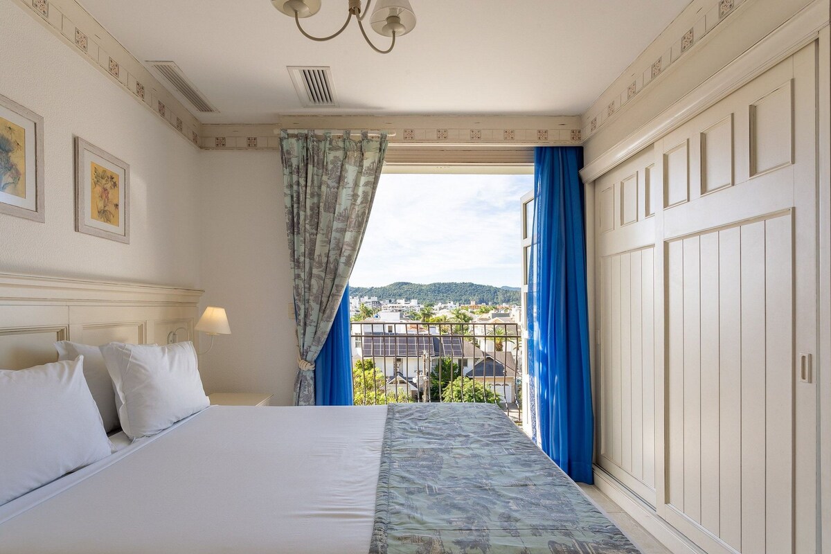 A hotel room with a bed, a nightstand, a lamp, and a view of the city through a window with blue curtains.