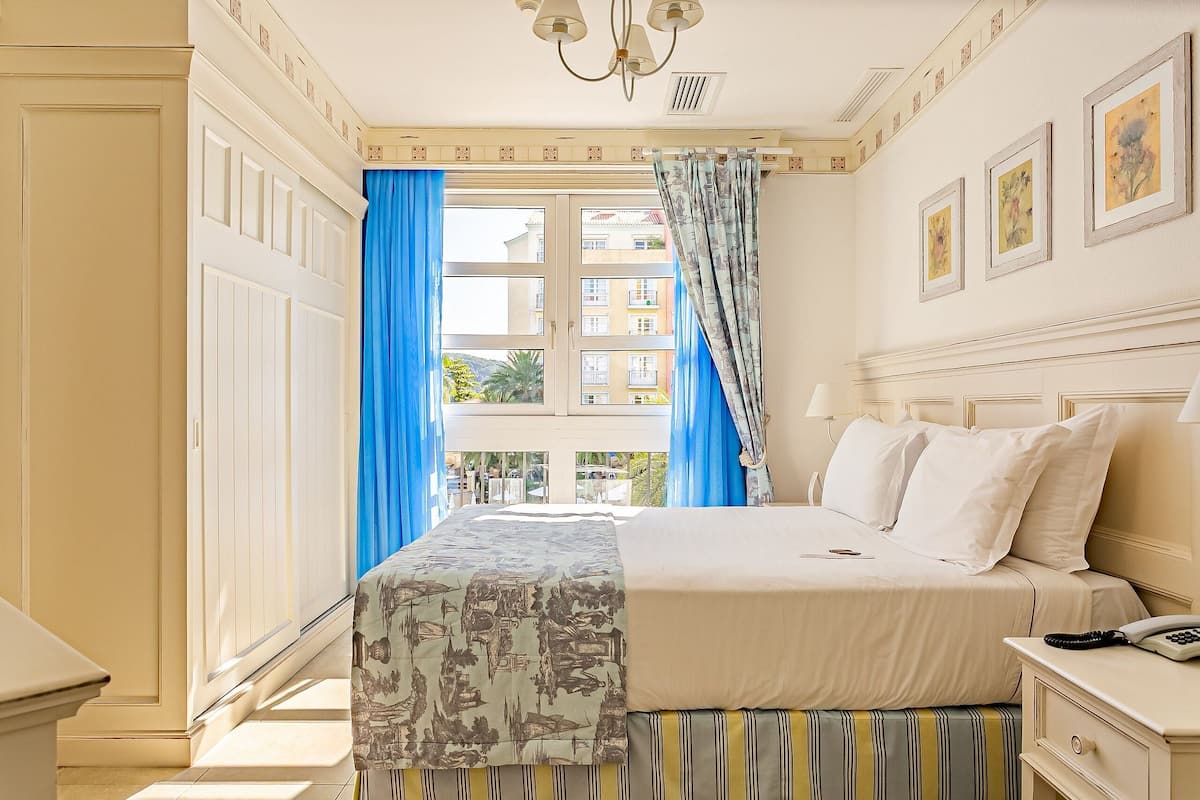 Una habitación de hotel con cama, mesita de noche, lámpara y ventana con cortinas azules.