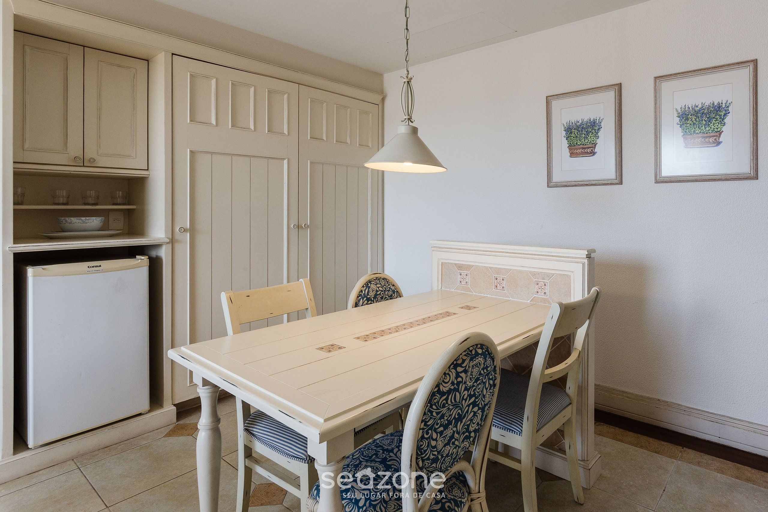 Un comedor con mesa y sillas blancas, una lámpara colgante y un refrigerador.