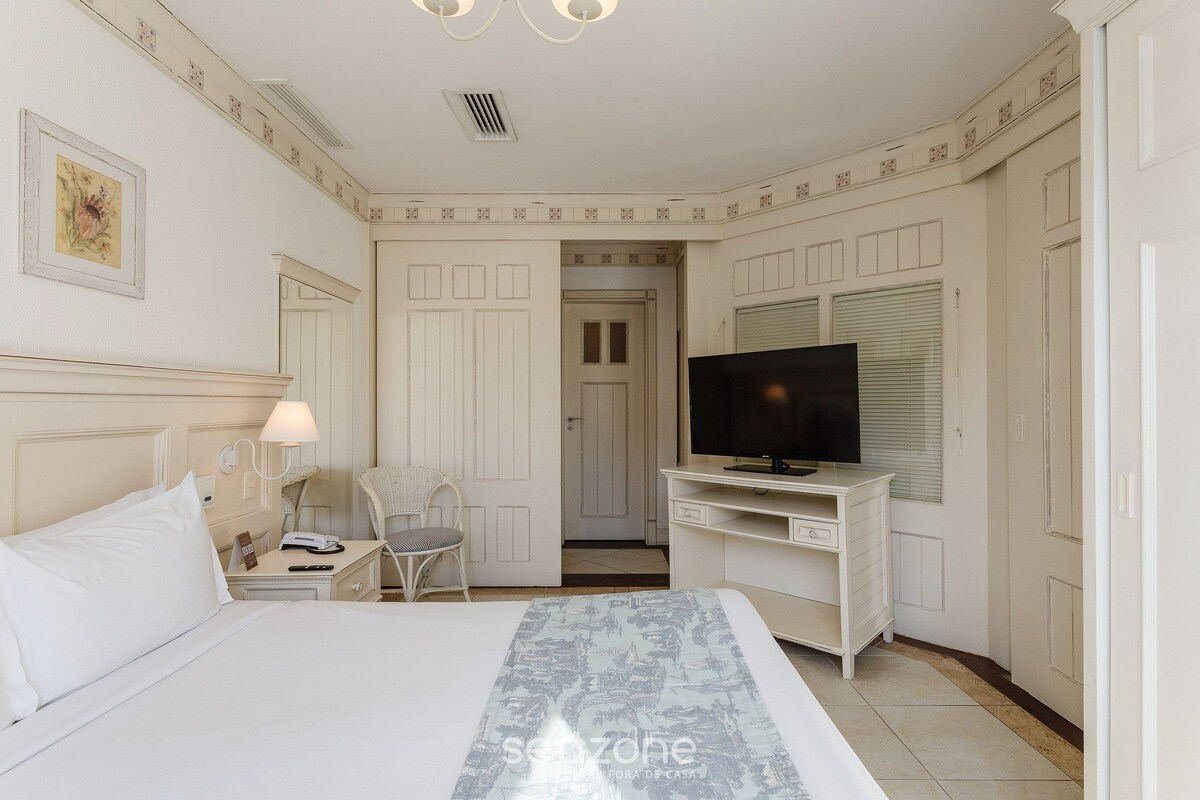 A hotel room with a bed, a desk with a TV, a chair, and a lamp.