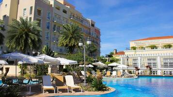 A hotel pool area with multiple lounge chairs and umbrellas, surrounded by palm trees and a large building in the background.