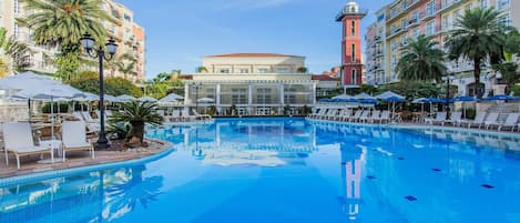 A swimming pool with lounge chairs and umbrellas, surrounded by multi-story buildings with balconies and palm trees.
