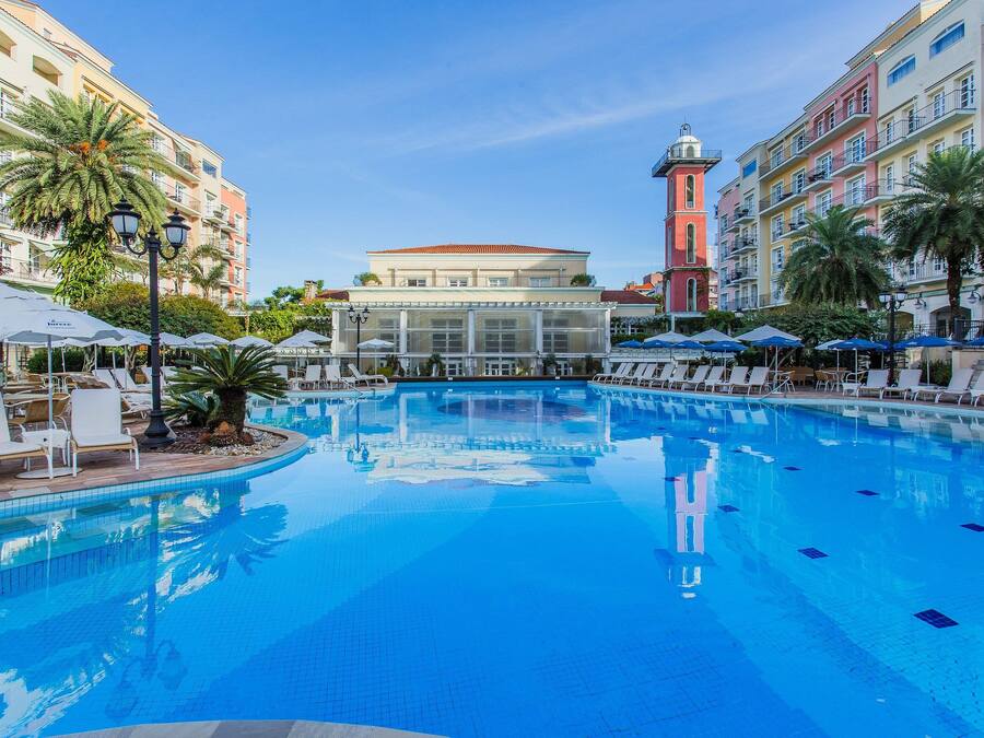 Una piscina con sillas de descanso y sombrillas, rodeada de edificios de varios pisos con balcones y palmeras.