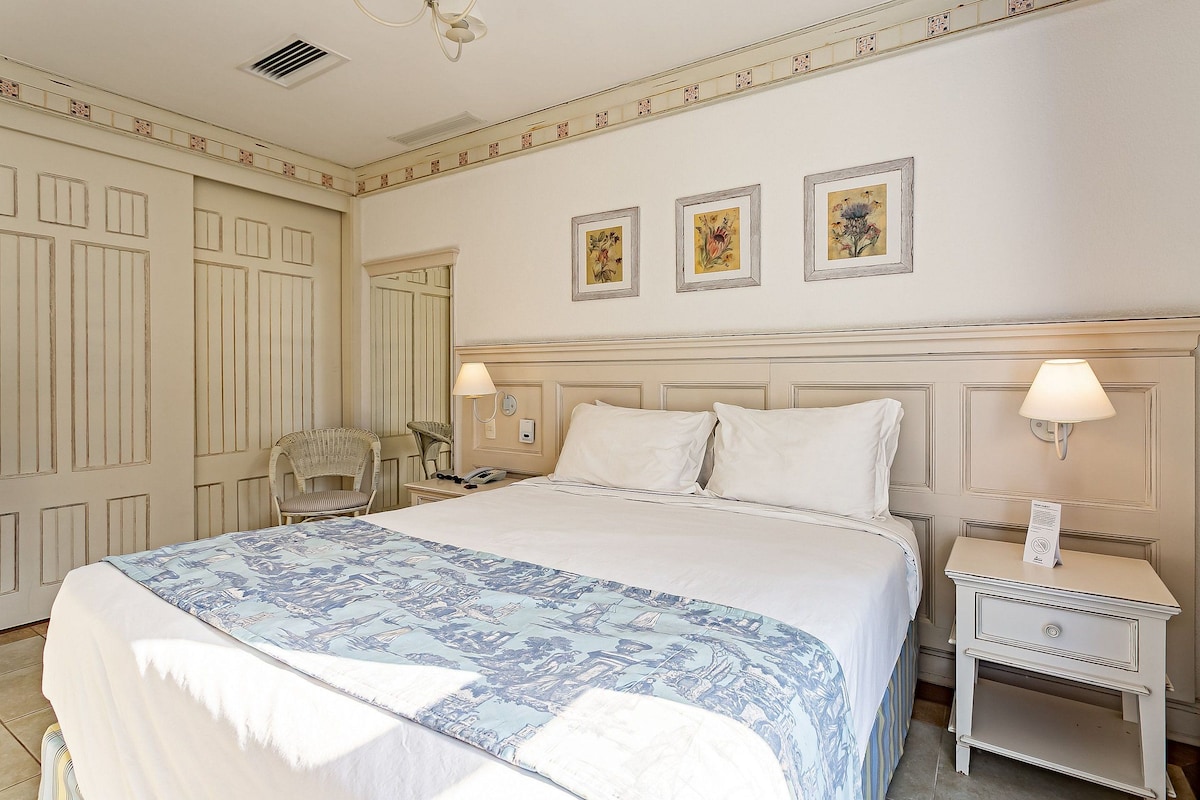 Una habitación de hotel ordenada con una cama grande, dos lámparas de noche, una mesita de noche y una silla.