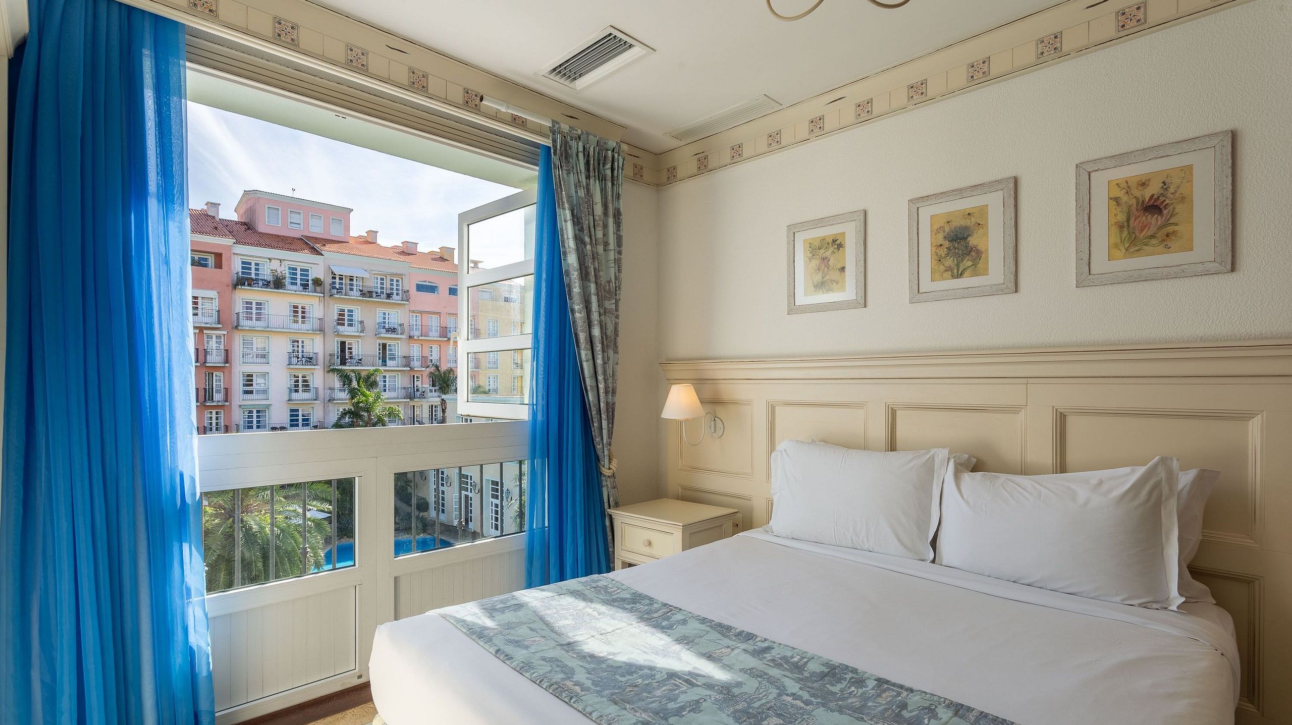 Habitación de hotel con una cama grande, una mesita de noche, un ventanal con cortinas azules y tres cuadros enmarcados en la pared.