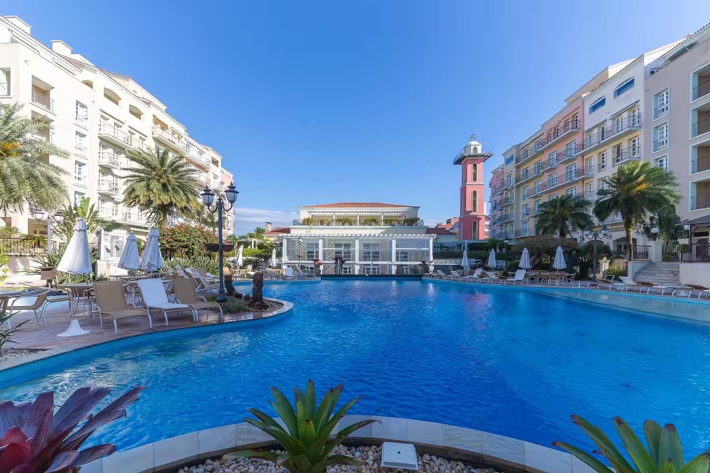 A swimming pool surrounded by lounge chairs and umbrellas, with a building featuring a tower in the background.