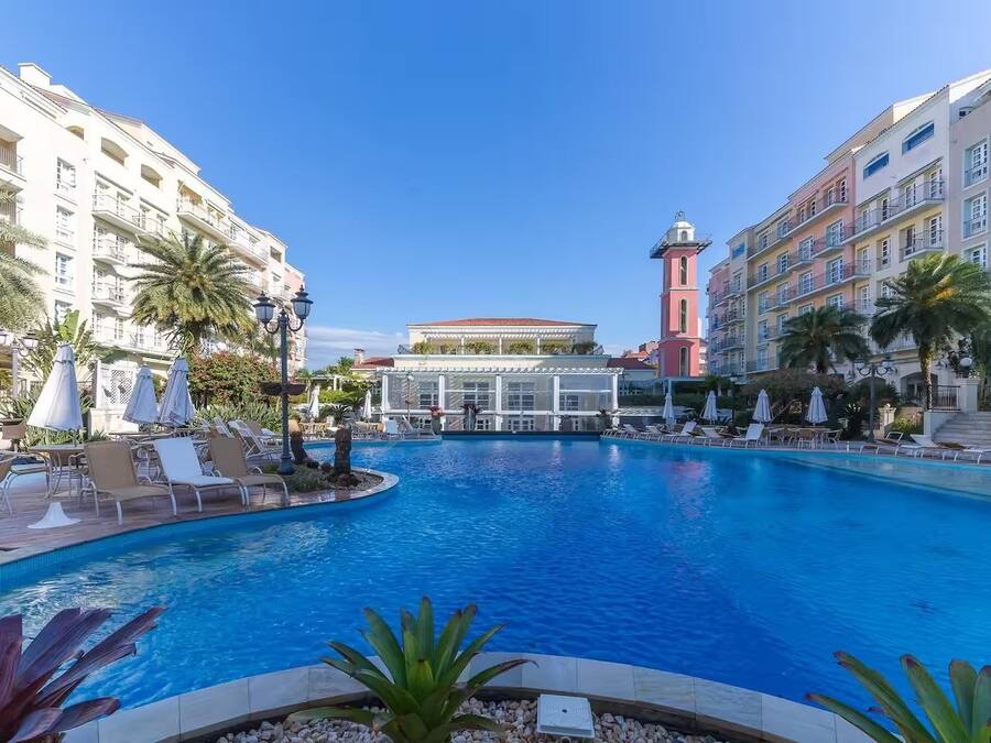 Piscina rodeada por espreguiçadeiras e guarda-sóis, com um edifício e uma torre ao fundo.